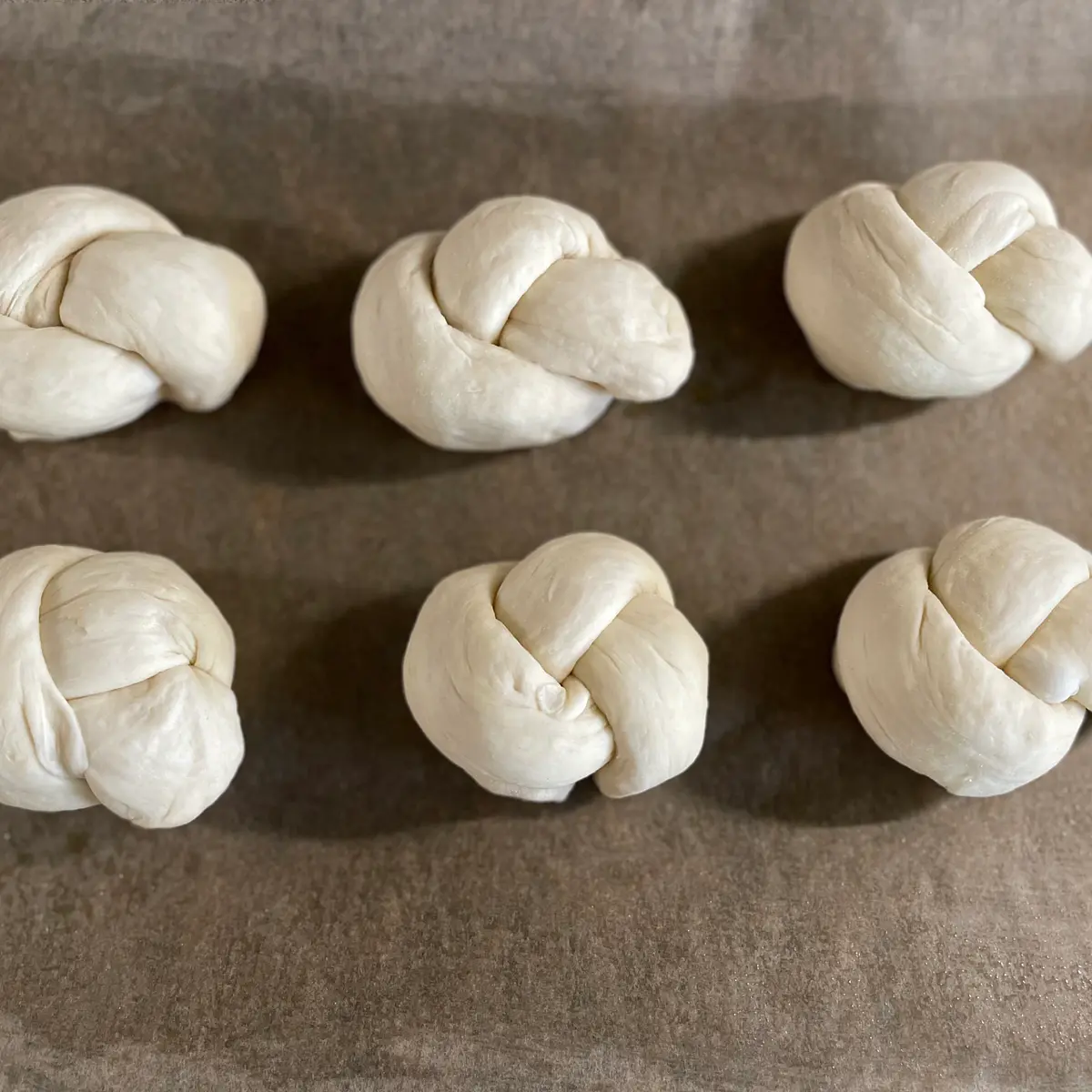 Divide the dough into 12 blobs – you might find it helpful to use some scales here (they will probably need to weigh around 80g each, although this may vary a bit). For 10 larger rolls, they will be about 95g each (of course, you could make 8 even larger rolls! Up to you.) Roll each blob into a long-ish “sausage”, about 20 cm long. Take each sausage of dough, tie it into a knot (everyone has their own style!), and place them on the tray, leaving a few cm between each one. As they prove again, they will expand; having them touch each other when cooking is fine (and it’s quite nice to tear them apart and have little soft edges), but you don’t want them touching before the second prove as they’ll be too squashed together).