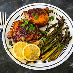 A delicious home-cooked meal featuring crispy, pan-seared chicken thighs garnished with fresh flat-leaf parsley, served alongside tender roasted asparagus spears and fresh lemon slices.