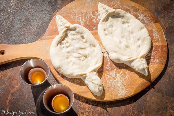 Prepare everything you need to shape and bake Khachapuri. Using BBQ/grill silicone mats makes the process easier. Dust the mat with some flour. Take one portion of dough and stretch it into an oval using your hands. Pinch the opposite sides of the oval, as shown in the picture, to shape "handles."