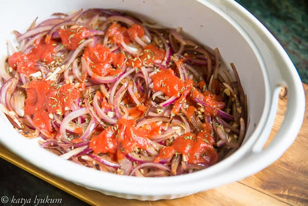 Transfer sauteed onions into the baking dish. Open a can of tomatoes, strain whole tomatoes, slice them in half lengthwise, and reserve. Top sauteed onions with a few spoons of tomato puree from the can. Sprinkle with minced herbs.