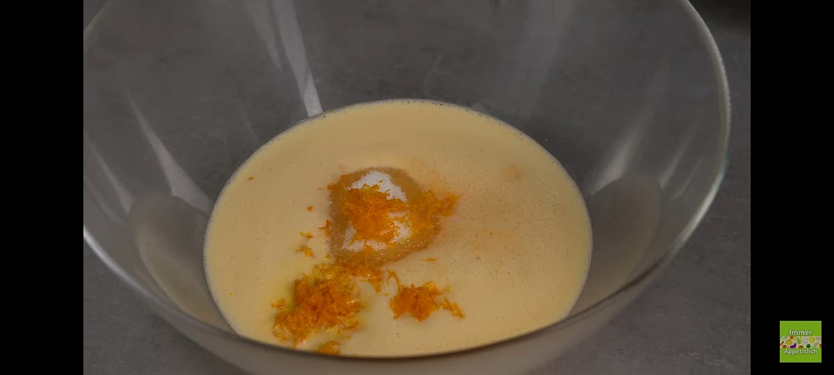 Añadimos al bol 100 gramos de azúcar y la piel de la mandarina que rallamos al principio y removemos con una batidora manual. Añadimos el aceite y el agua y removemos. Tamizamos 220 g de harina y 10 g de levadura y removemos.