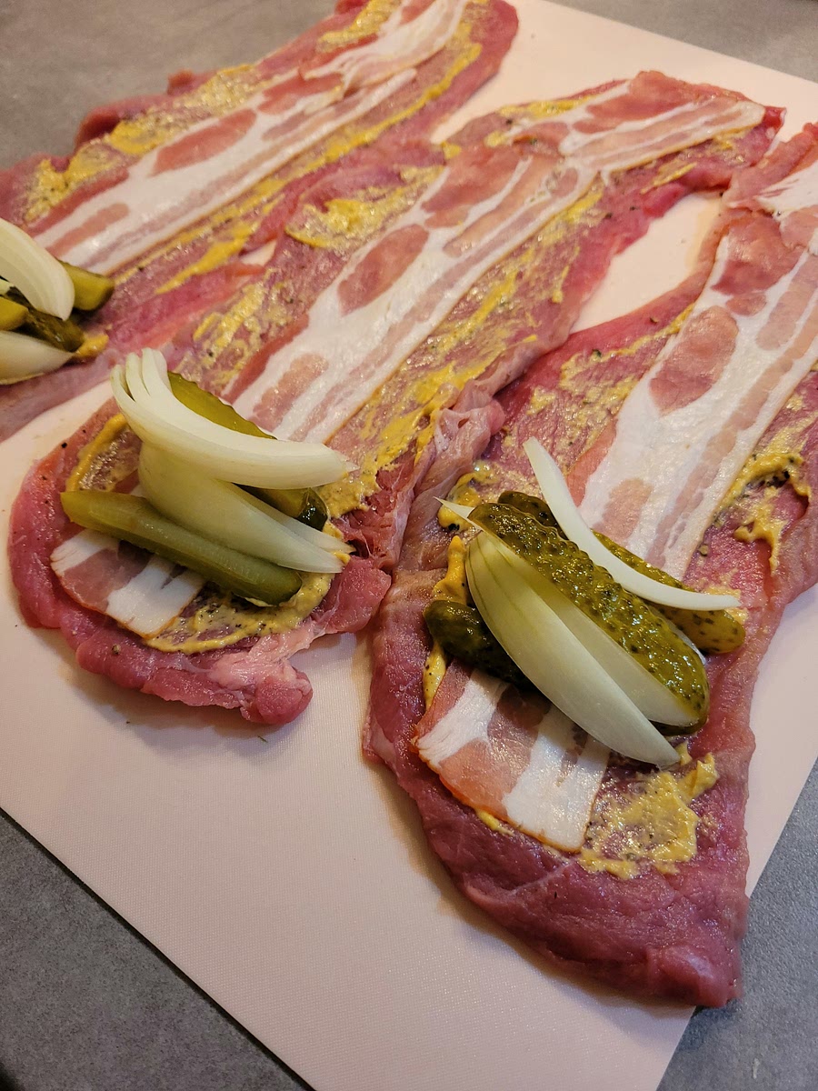 Die Rouladen flach ausbreiten, mit Pfeffer und Salz bestreuen und flächig dünn mit Senf bestreichen. Eine Scheibe Bacon auf jede Rouladen legen. Darauf die Zwiebelstücke und die Gurkenstreifen platzieren. Dann von der schmaleren Seite her aufrollen und mit Rouladennadeln feststecken.
