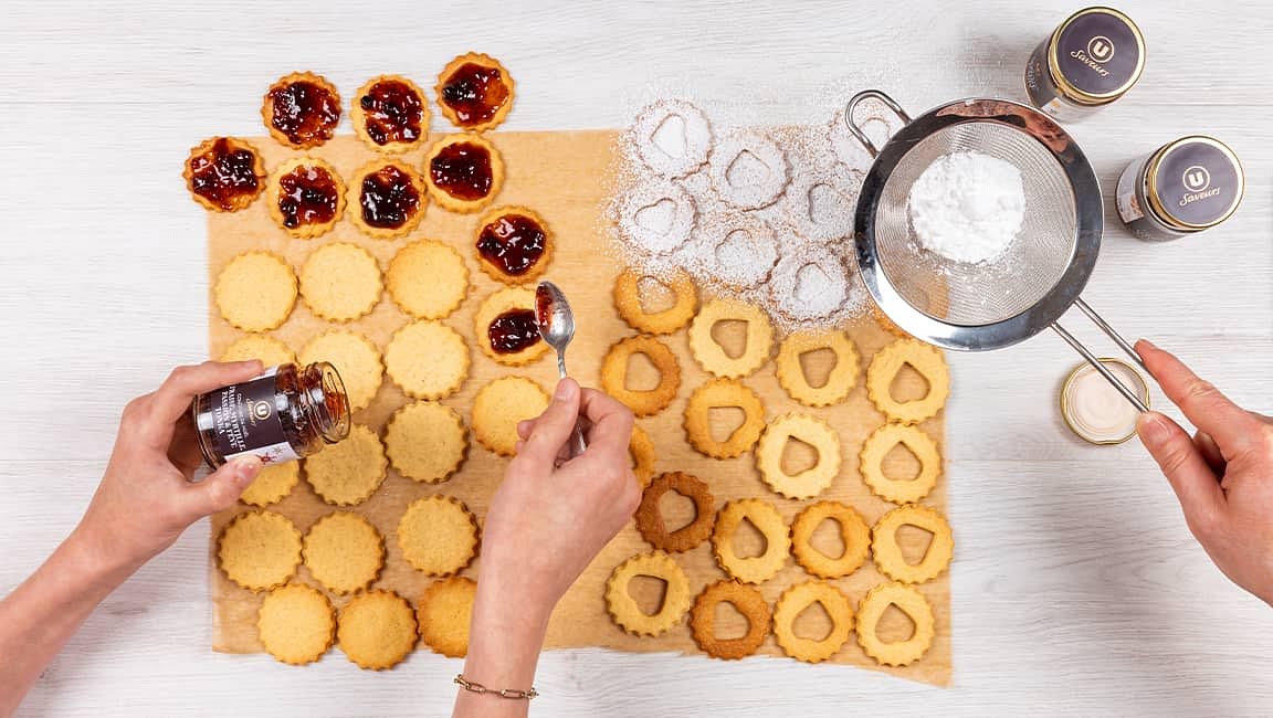 Enfournez les biscuits sur la plaque du four pendant 10 min à 180 °C. Après refroidissement, versez un peu de sucre glace dans la passoire et saupoudrez-en les biscuits troués. Étalez un peu de confiture sur les sablés entiers, puis posez les sablés troués dessus.