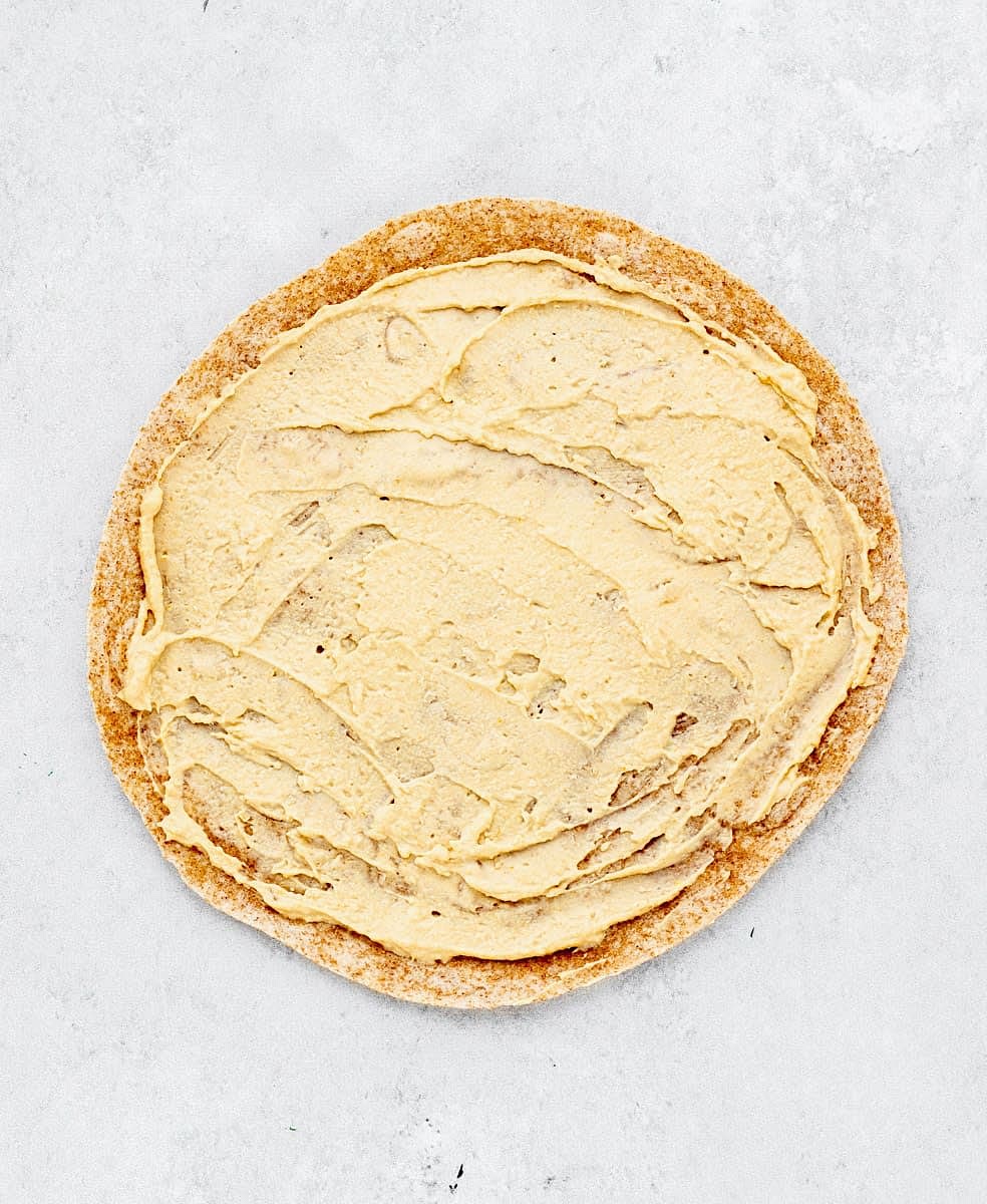 To start, spread a thin layer of hummus all over a large leaving about an inch around the edge, to add some flavour and act as a glue to keep all of the ingredients in place.