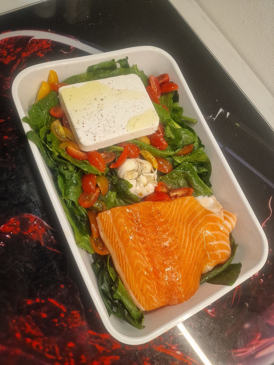 Als erstes bereiten wir die Auflaufform für den Ofenkäse vor. Der Backofen wird auf Ober-/Unterhitze auf 200 Grad vorgeheizt. Wir befüllen die Auflaufform: Wir schneiden den Kopf der Knoblauchknolle ab und legen diese in die Mitte. Drum herum drapiert wir den Spinat & die Cherrytomaten. Auf die eine Seite legen wir den Lachs auf die Haut, auf die andere Seite den Feta. Wir verteilen das aolivenöl, den Pfeffer und das Salz drüber. Dann geht die Auflaufform für 15 Minuten in den vorgeheizten Ofen.