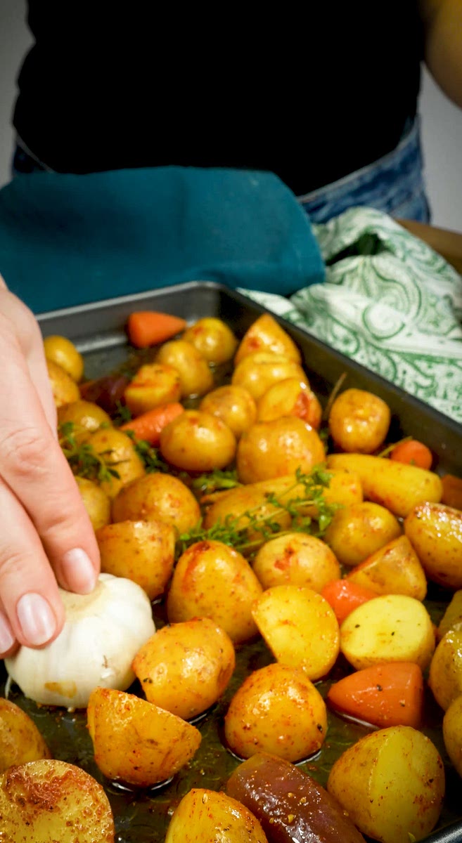 3. Add butter and 1 tablespoon of olive oil to the baking tray, then add the drained potatoes and carrots, season with salt, pepper, and paprika, and place the thyme springs on top. Mix well and add the garlic bulb (cut side down) to the tray.