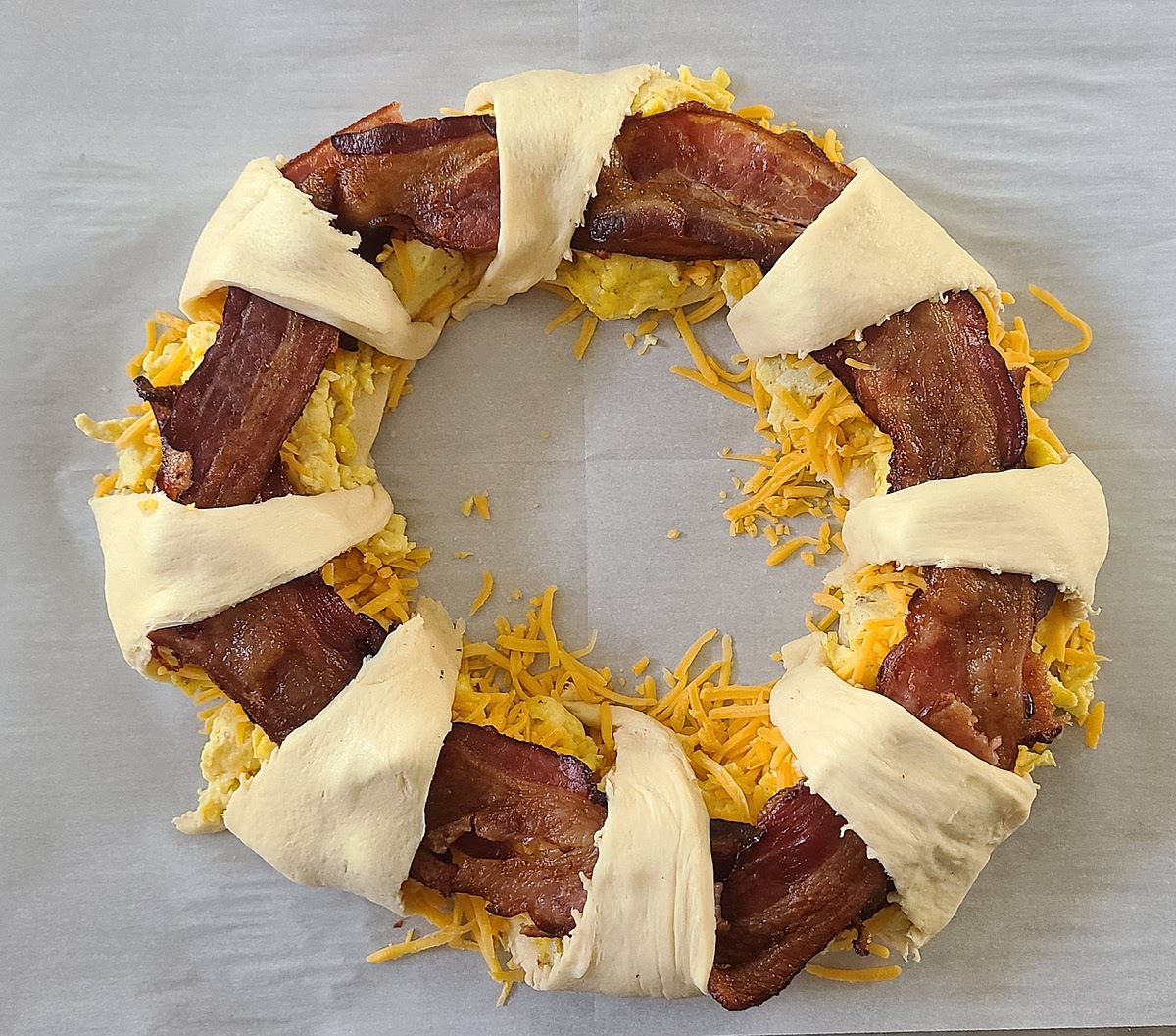 When bacon and eggs are done - Arrange the croissant dough triangles in a circle on a parchment lined baking tray, with the base of the triangles just barely overlapping. Layer the eggs in a circle on the croissant. Sprinkle the eggs generously with cheese (I never measure it). Then, top the mound with bacon. (I usually tear them in half and place one half per each croissant if using 4 pieces of bacon, then overlap them again between the croissants if using 8.) Finally, tuck the small end of the croissant dough over the egg/cheese/bacon mound and under the base side of the croissant dough.