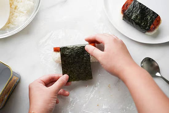 Carefully wrap the nori around the Spam musubi. I like to lightly press the nori on the sides and then lift the top to slide in the nori underneath and repeat for the other side.