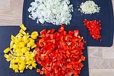 Die Zwiebeln und die Knoblauchzehen schälst Du und schneidest sie in kleine Würfel. Die Peperoni waschen, entkernen, und ebenfalls in kleine Würfel schneiden. Die rote(n) und gelbe(n) Paprikaschoten waschen, entkernen und in mundgerechte Stücke schneiden.