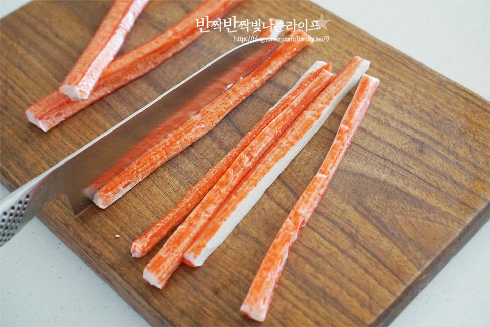 생선에 게향을 첨가하여 만든 게맛살은 아이들반찬으로 빠지지 않는 재료인데요. 김밥이나 샐러드와 같은 요리에 넣어 먹기도 하는데 오늘은 하트모양으로 만들어 전을 구워줄 것이랍니다. 먼저 개별 포장된 패키지를 벗긴 뒤 세로 길이대로 길게 반으로 잘라줍니다. 큼직하게 맛보고 싶다면 그대로 구워주셔도 좋습니다. 양 끝부분을 잡고 가운데로 동그랗게 말아 넣어 하트로 모양을 내어줍니다. 그리고 나무꼬치나 이쑤시개를 가운데로 꽂아 풀리지 않게 고정해 줍니다. 이 때 평평한 바닥면에 올려 고정해 주면 안에 들어갈 계란물이 새어 나오지 않고 예쁘게 굽기 좋습니다.