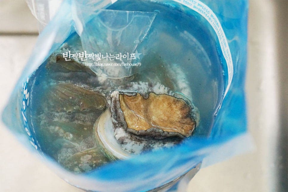 전복회 손질을 위해 살아있는 생물을 준비했습니다. 바로 먹는 것이다 보니 신선도가 중요해요. 완도 보길도 산지에서 산소포장하여 살아서 움직이는 게 싱싱해요. 4마리만 회로 썰기 위해 준비합니다. 적당히 큼직하고 집었을 때 움직이며 탄력이 있는 것이 신선함 가득 집에서 먹기 딱 좋아요. 조리용 솔을 준비합니다. 칫솔도 좋습니다. 솔로 문질러가며 씻어준 뒤 흐르는 물에 헹구어 줍니다. 너무 세게 문지르게 되면 오히려 쫄깃한 식감이 물러질 수 있으니 조심하여 씻어주세요. 살을 썰어서 껍질 위에 담아줄 것이라 껍질도 문질러 씻어줍니다. 씻어준 뒤 껍질에서 살을 분리해 주는데요. 동그랗게 뚫린 뾰족한 게 입이에요. 입쪽으로 숟가락을 넣어 분리해 줍니다. 내장이 터지지 않도록 안쪽 붙어 있는 쪽을 밀어내면 쉽게 분리가 됩니다. 적은 양의 끓는 물에 껍질이 닿도록 10초 이내로 살짝 데쳐서 분리해 주셔도 좋아요. 살과 내장이 슥 떨어지면 내장과 살도 떼어내어 줍니다. 이 과정에서 주의하실 점은 터지지 않도록 해주셔야 해요. 한두 개 하다보면 요령이 생긴답니다~ 뒤집어보면 검은색 입에 이빨이 있어요. 전복이빨제거를 위해 살짝 눌러주거나 칼로 저미듯이 썰어주시면 아래 사진과 같이 깔끔하게 손질됩니다. 내장에 보면 볼록하게 튀어나온게 모래주머니인데요. 내장 위쪽(손가락이 위치한 방향)을 보면 타원형 모양으로 길게 붙은 게 모래주머니에요. 가위로 잘라 제거한 뒤 드시면 고소하고 식감도 좋습니다. 내장은 좋아하지 않아 냉동 소분하여 죽에 넣곤 하는데요. 끓는 물에 살짝 데쳐서 곁들이면 되어요. 흐르는 물에 깔끔하게 헹구어 키친타월로 물기를 제거해 줍니다.