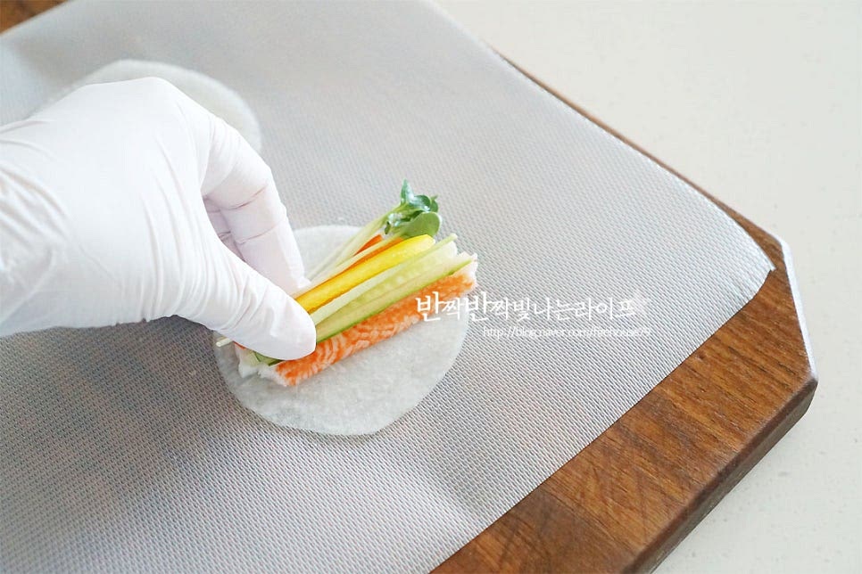 쌈무를 먼저 펼친 뒤 그 위로 재료를 조금씩 올려주는데요. 속재료를 너무 많이 넣으면 말이 모양이 잡히지 않으니 종류별로 조금씩 올려주시면 됩니다. 양 끝 부분을 부채꼴 모양으로 감싸 말아줍니다. 얇게 썬 절임무라서 그대로 모양을 잡아주어도 고정이 되지만 색감도 더해주고 고정해주기 위해 부추로 묶어줍니다. 색감이 겹치지 않게 재료를 넣어 말아주시면 만들었을 때 먹음직스러워요.