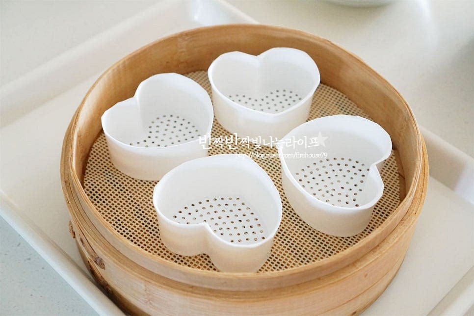 찜기에 면포를 깔고 하트틀을 올려줍니다. 사각틀이나 머핀 틀 등 다양한 도구를 활용해 주면 됩니다. 만들어둔 백설기 반죽을 넣어줍니다. 이때 꾹꾹 눌러 담지 않고 솔솔 뿌려가며 자연스럽게 올려주세요. 사이사이 공기가 들어가면서 부드럽고 맛있게 쪄진답니다. 두 가지 색을 각각 담은 뒤 스크레이퍼로 평평하게 정돈해 줍니다. 이때도 눌러 담지 않고 자연스럽게 정돈해 주세요.