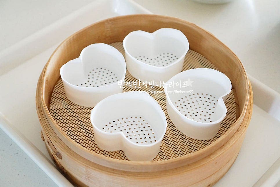 찜기에 면포를 깔고 하트틀을 올려줍니다. 사각틀이나 머핀 틀 등 다양한 도구를 활용해 주면 됩니다. 만들어둔 백설기 반죽을 넣어줍니다. 이때 꾹꾹 눌러 담지 않고 솔솔 뿌려가며 자연스럽게 올려주세요. 사이사이 공기가 들어가면서 부드럽고 맛있게 쪄진답니다. 두 가지 색을 각각 담은 뒤 스크레이퍼로 평평하게 정돈해 줍니다. 이때도 눌러 담지 않고 자연스럽게 정돈해 주세요.