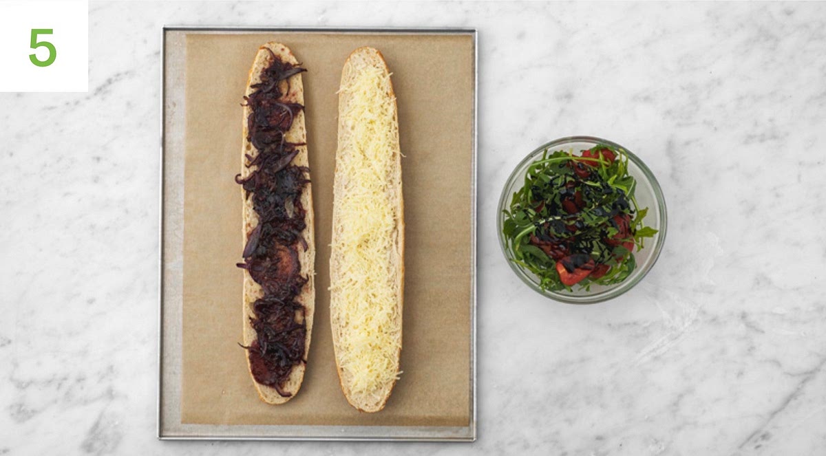 While the steaks cook, slice the baguette in half lengthways. Spread the caramelised onions over the base and sprinkle the Cheddar over the lid. Return the baguette to the middle shelf of the oven until the cheese has melted, 3-5 mins. Meanwhile, when the steaks are rested, slice widthways into 1cm slices. Once the cheese has melted, remove the baguette from the oven. Top with the sliced steak and spoon over 2 tbsp of the red wine jus, then sandwich shut and slice in half widthways. Just before everything's ready, in a large bowl, toss the rocket and tomatoes with the balsamic glaze.