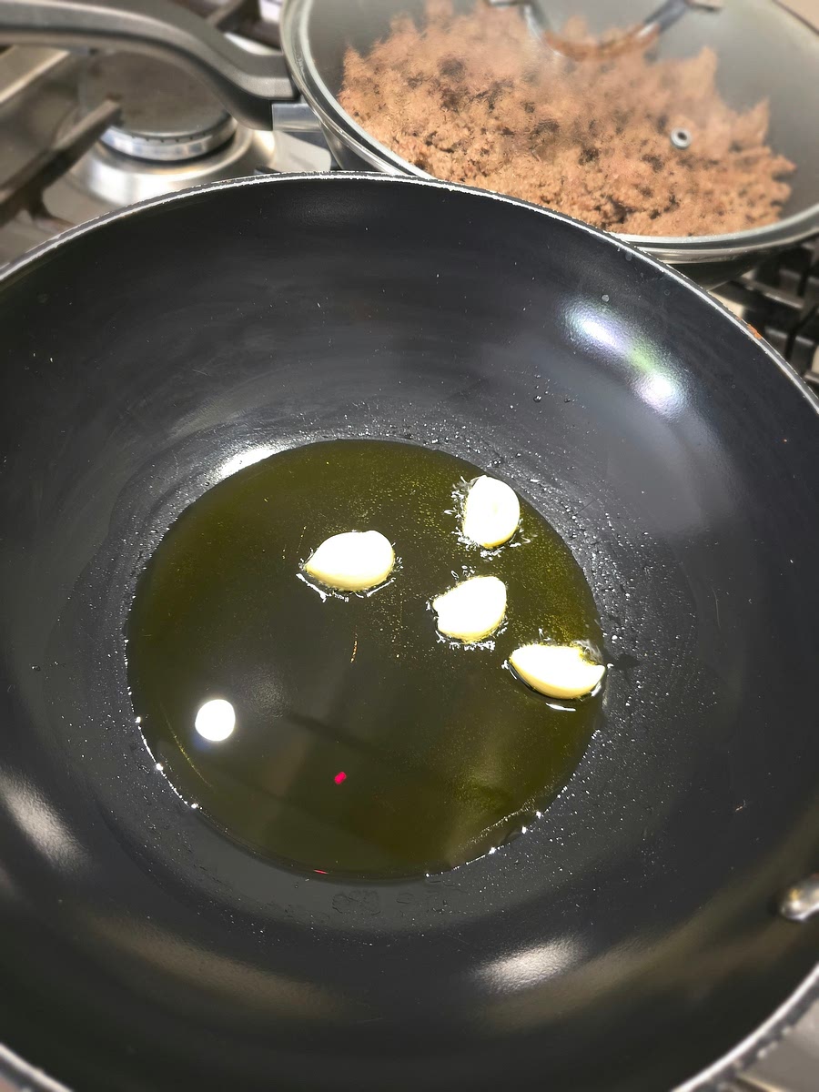 Doe 5 a 6 el olijfolie in een grote pan (wok) en leg de knoflooktenen er in. Laat lekker bakken tot de knoflook een beetje bruin wordt en zacht. Zet het vuur niet te hoog en draai met een tang of vork de knoflook om anders loop je de kans dat het zwart wordt en moet je opnieuw beginnen. Leg daarna de knoflook op een houten snijplank, bestrooi met flink wat zout en prak met een vork. Daarna mag de geprakte knoflook weer terug in de olie. Dit is de basis voor je saus!