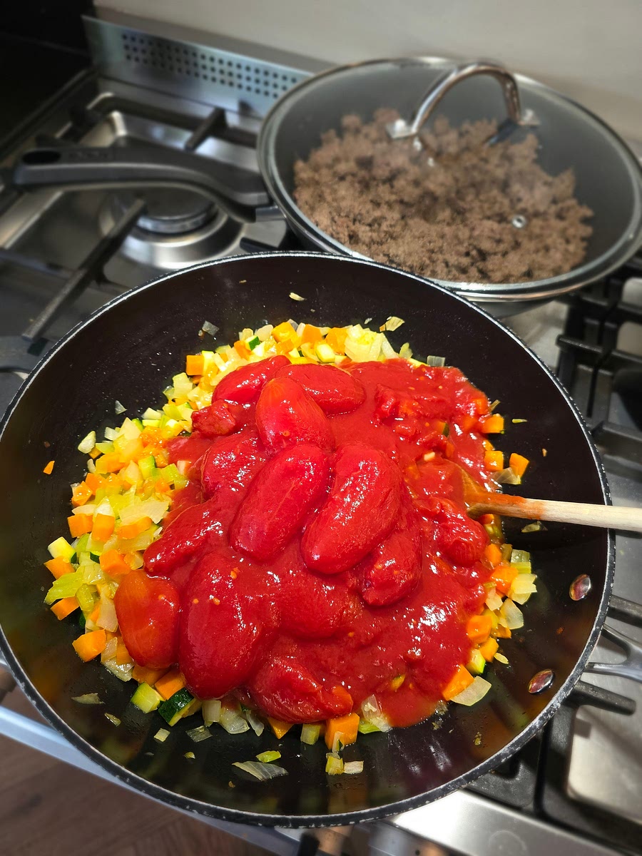 Als de groenten voldoende zacht zijn is het tijd om de blikken tomaat er bij te voegen. Je kunt elke variatie gebruiken. In dit geval heb ik drie blikken gepelde tomaten en twee blikken tomatenblokjes met knoflook. Ik had nog ergens drie tomaten over, die heb ik er ook doorheen gedaan. Goed roeren en peper en eventueel zout toevoegen en opnieuw roeren. Nu kun je een warmhoudplaatje tussen de pan en het vuur zetten, mocht je ook op gas koken. Het is nu 14:15 en dus ruim voldoende op de saus lekker op te laten staan.