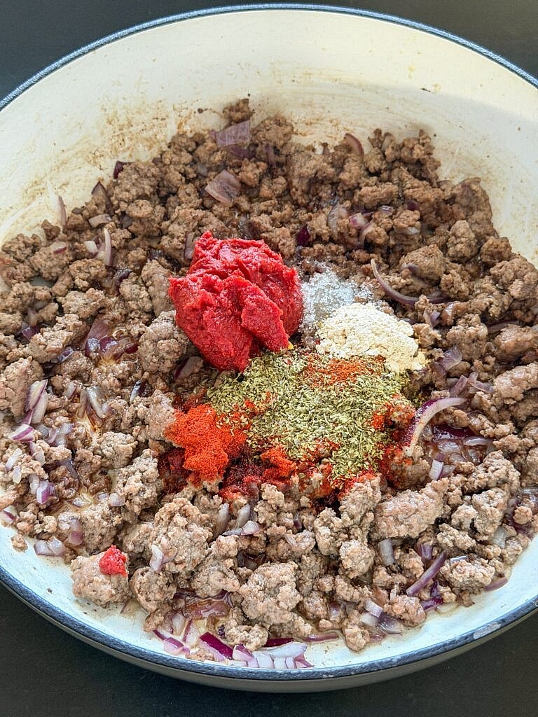 Voeg de tomatenpuree, 1 theelepel uienpoeder, 1,5 eetlepel gedroogde oregano , 1/3 theelepel chilivlokken, 1/2 theelepel gerookte paprikapoeder, 2 theelepels paprikapoeder en 1/2 theelepel suiker en knoflook toe. Bak dit ongeveer 30 seconden mee zodat de kruiden en tomatenpuree meer smaak krijgen.